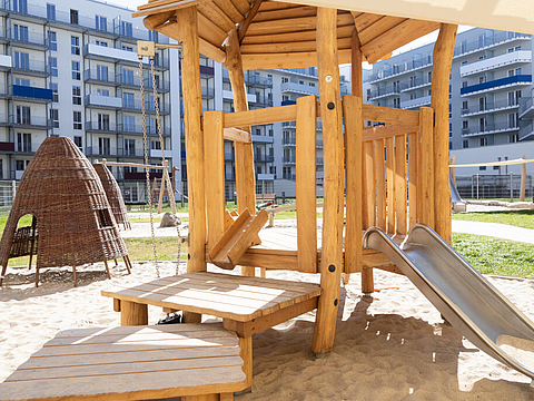 Es gibt einen Kletterturm aus Holz mit Dach und Rutsche und Geräte, die dazu einladen mit Sand zu bauen und zu spielen.